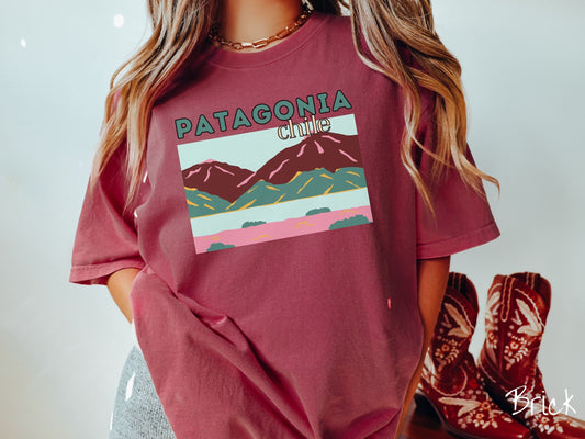 A woman wearing a cute, vintage brick colored Comfort Colors t-shirt with the text Patagonia Chile in green and yellow font. Below this is a picture of Patagonia with brown and green mountains, a river, and pink sandy beaches with green bushes.