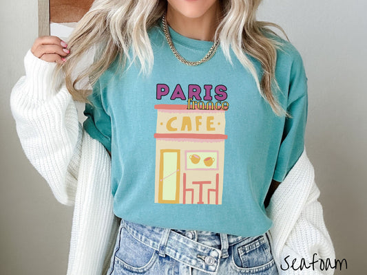 A woman wearing a cute, vintage seafoam colored Comfort Colors t-shirt with the text Paris France in purpple and yellow font. Below this is a French cafe with a coffee cup picture on the outside above a table and chairs for outside seating.