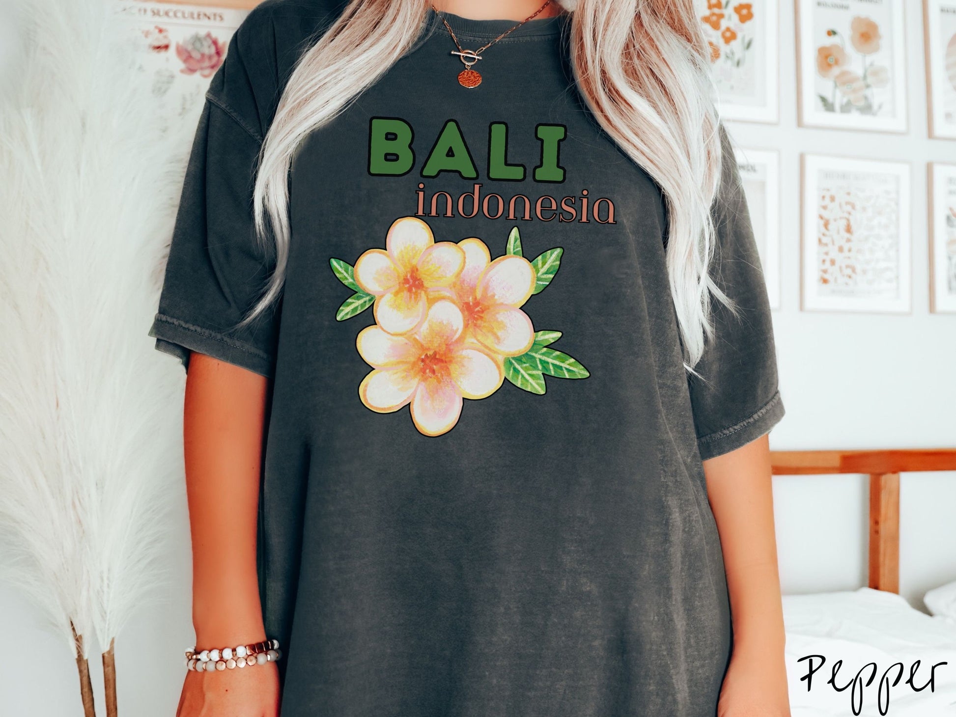 A woman wearing a vintage, pepper colored shirt with the text Bali Indonesia in green and yellow font, respectively, and a picture underneath of three beautiful yellow and orange flowers with green leaves.