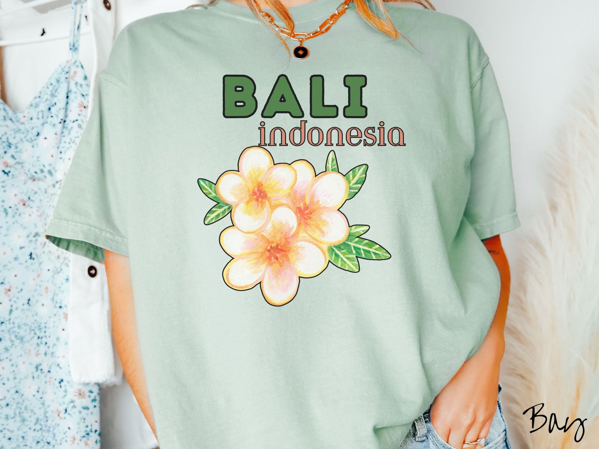 A woman wearing a vintage, bay colored shirt with the text Bali Indonesia in green and yellow font, respectively, and a picture underneath of three beautiful yellow and orange flowers with green leaves.