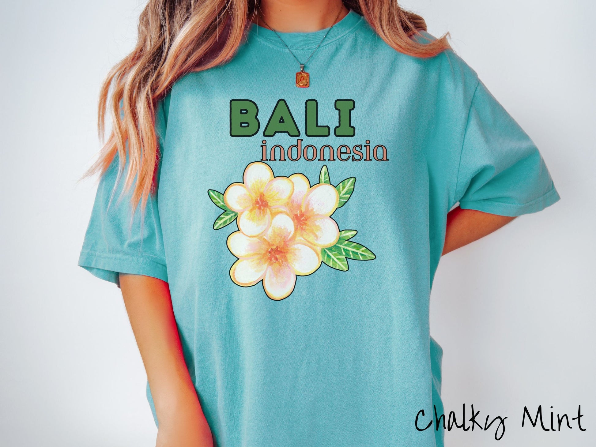 A woman wearing a vintage, chalky mint colored shirt with the text Bali Indonesia in green and yellow font, respectively, and a picture underneath of three beautiful yellow and orange flowers with green leaves.