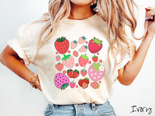 A woman wearing a cute, vintage ivory colored Comfort Colors t-shirt with many different strawberries displayed in a grid layout.There are red, pink, and various shades of strawberres scattered among red flowers.