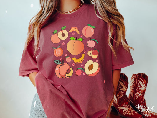 A woman wearing a cute, vintage brick colored Comfort Colors t-shirt with many different peaches displayed in a grid layout.There are whole peaches, halves of peaches, and quartes of peaches, with various pictures of the peach pit.