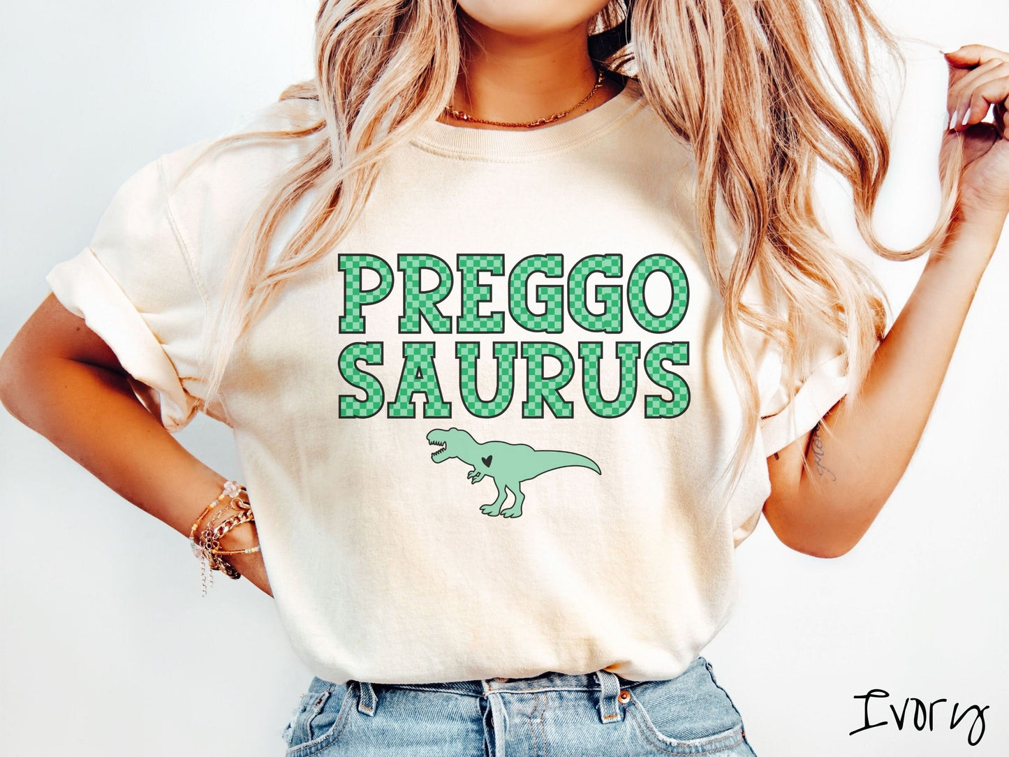 A woman wearing a vintage, ivory colored Comfort Colors t-shirt with the text PREGGO SAURUS in green-block, uppercase font. Below this is a green tyrannosaurus rex with its mouth open showing teeth and a black heart on its chest.