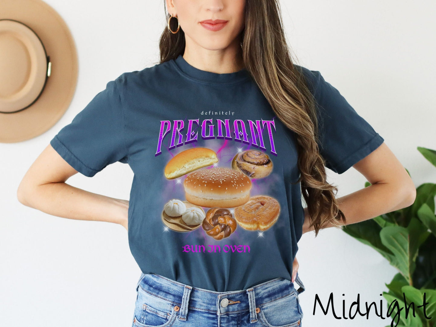 A woman wearing a vintage, midnight colored Comfort Colors t-shirt with the text Definitely Pregnant Bun In Oven in purple font. Between the text are different breads like hamburger and hotdog buns, cinnamon rolls, and donuts.