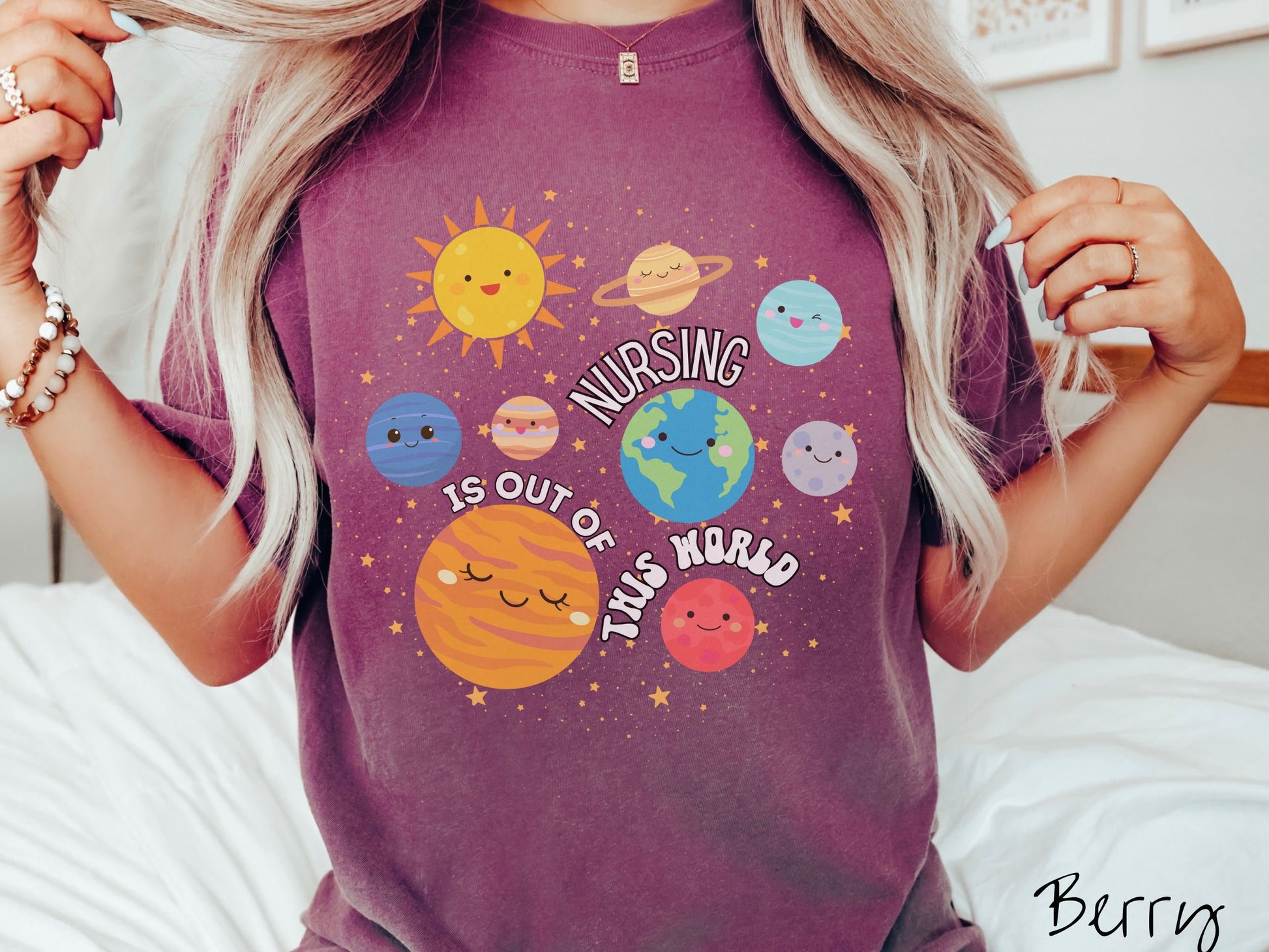 A woman wearing a cute, vintage berry colored shirt showing the solar system and all the planets have smiling, happy faces. There is text saying Nursing Is Out of This World among the planets, and there are many stars twinkling in the background.