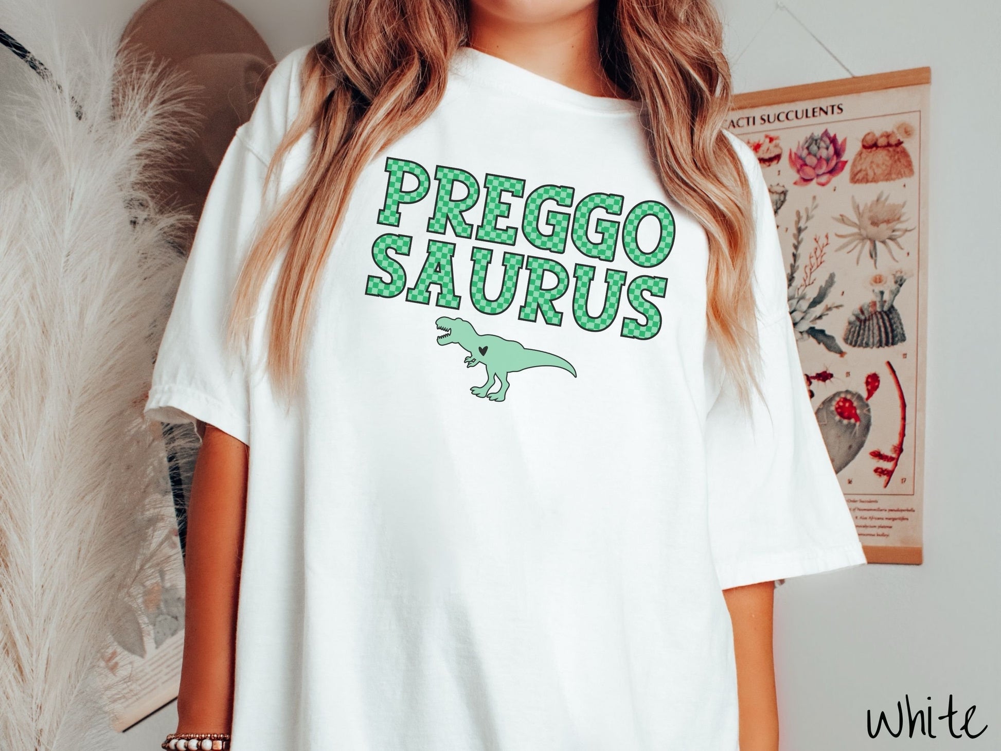 A woman wearing a vintage, white colored Comfort Colors t-shirt with the text PREGGO SAURUS in green-block, uppercase font. Below this is a green tyrannosaurus rex with its mouth open showing teeth and a black heart on its chest.