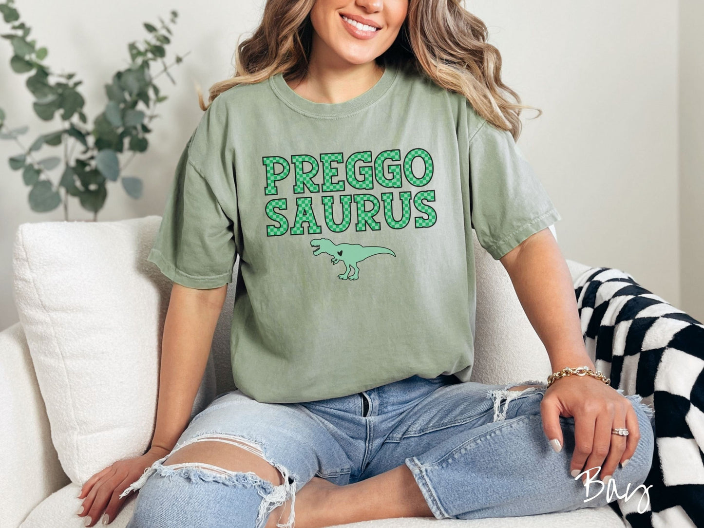 A woman wearing a vintage, bay colored Comfort Colors t-shirt with the text PREGGO SAURUS in green-block, uppercase font. Below this is a green tyrannosaurus rex with its mouth open showing teeth and a black heart on its chest.