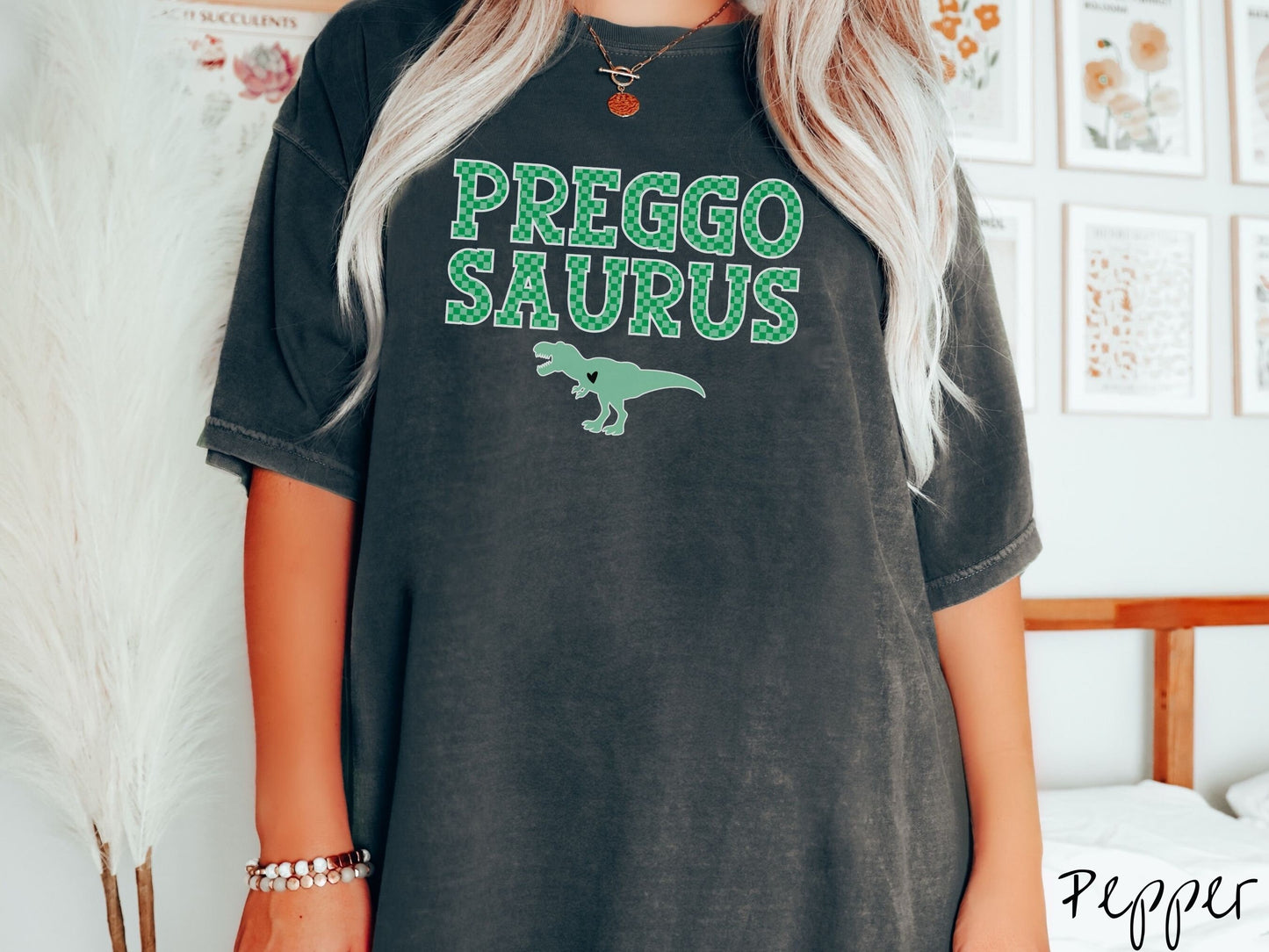 A woman wearing a vintage, pepper colored Comfort Colors t-shirt with the text PREGGO SAURUS in green-block, uppercase font. Below this is a green tyrannosaurus rex with its mouth open showing teeth and a black heart on its chest.
