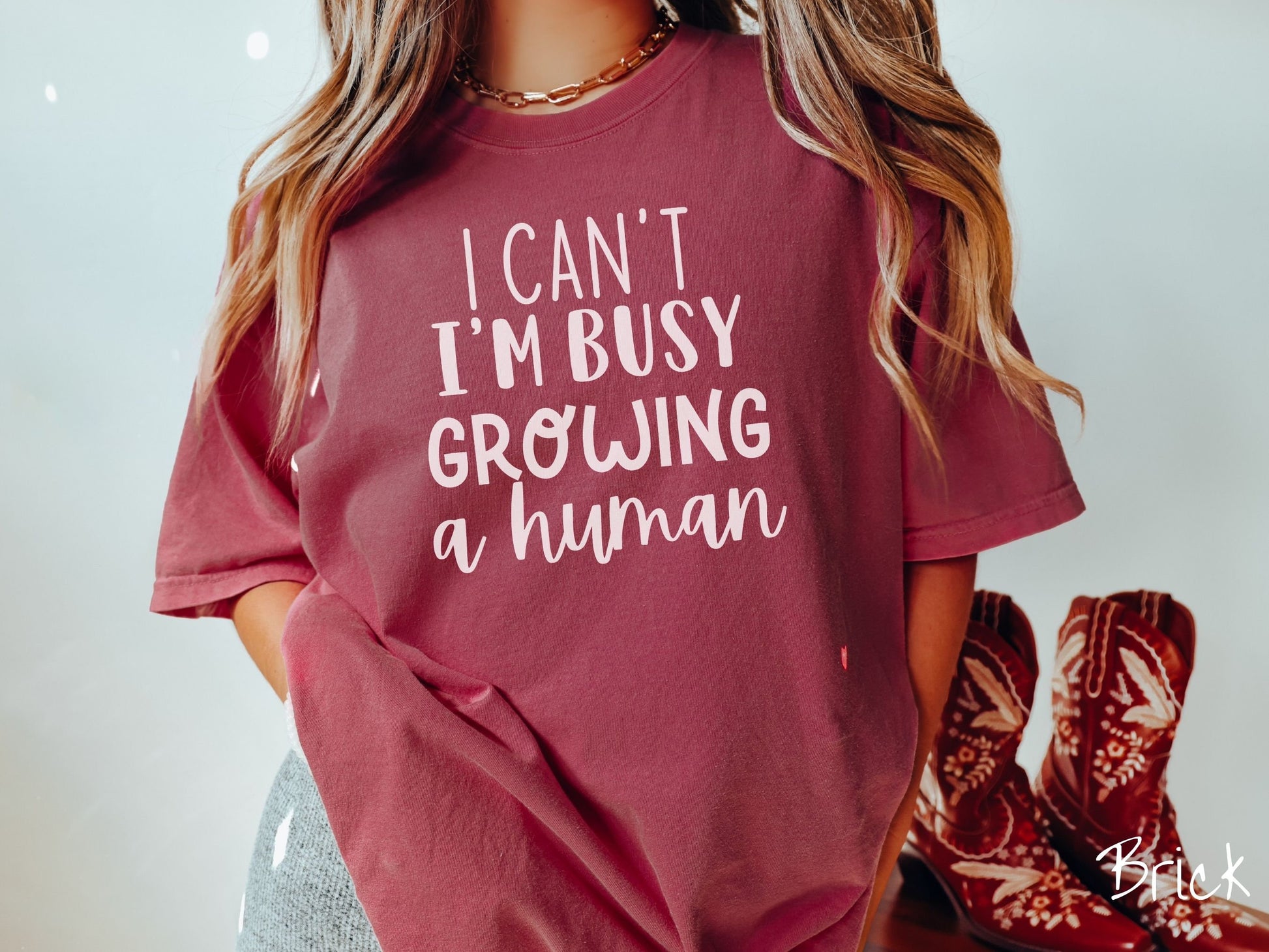 A woman wearing a vintage, brick colored Comfort Colors t-shirt with the text in white font I Can’t I’m Busy Growing A Human.
