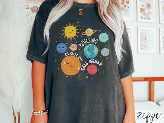 A woman wearing a cute, vintage pepper colored shirt showing the solar system and all the planets have smiling, happy faces. There is text saying Nursing Is Out of This World among the planets, and there are many stars twinkling in the background.