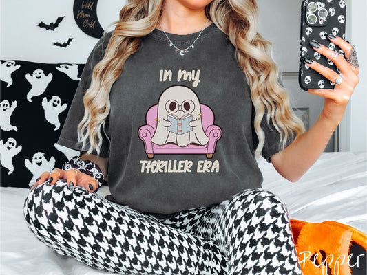 A woman wearing a vintage, pepper colored Comfort Colors t-shirt with the text In My Thriller Era across the front in ivory font. In between the text is a young ghost with glasses reading a book and sitting in a pink chair.