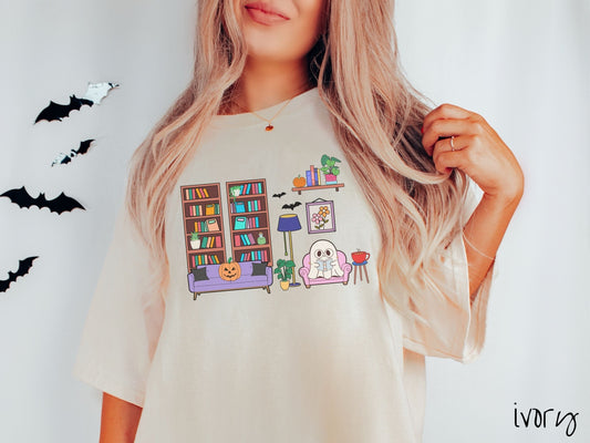 A woman wearing a vintage, ivory colored Comfort Colors t-shirt with a cozy Halloween-themed room. A young white ghost is reading a book in a chair next to a coffee and colorful bookcase. An orange pumpkin sits on the couch and bats are in the air.