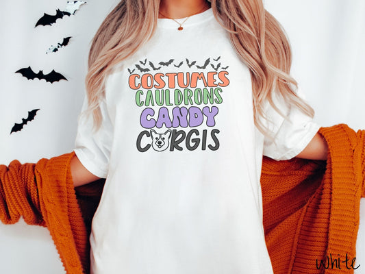 A woman wearing a vintage, white colored Comfort Colors t-shirt with the text listed vertically Costumes Cauldrons Candy Corgis in orange, green, purple, and black font. The O in Corgis is a smiling dog face. Above all the text are black bats.