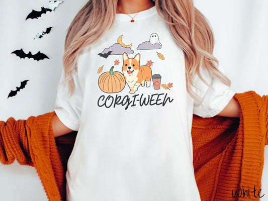 A woman wearing a vintage, white colored Comfort Colors t-shirt with the text Corgi-Ween across the front in black, cursive font. Above are a smiling corgi dog, an orange pumpkin, a pumpkin-spiced latte, and fall leaves, a ghost, and bat in the air.