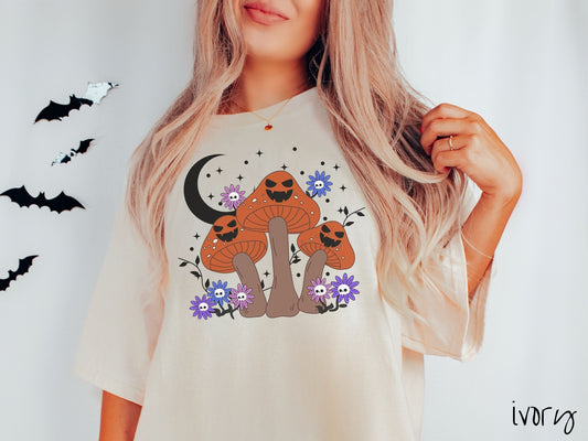 A woman wearing a cute, vintage ivory colored Comfort Colors T-shirt with three dark orange and light brown mushrooms with black jack o&#39;lantern faces. They are standing around blue and purple flowers and in front of a black crescent moon.