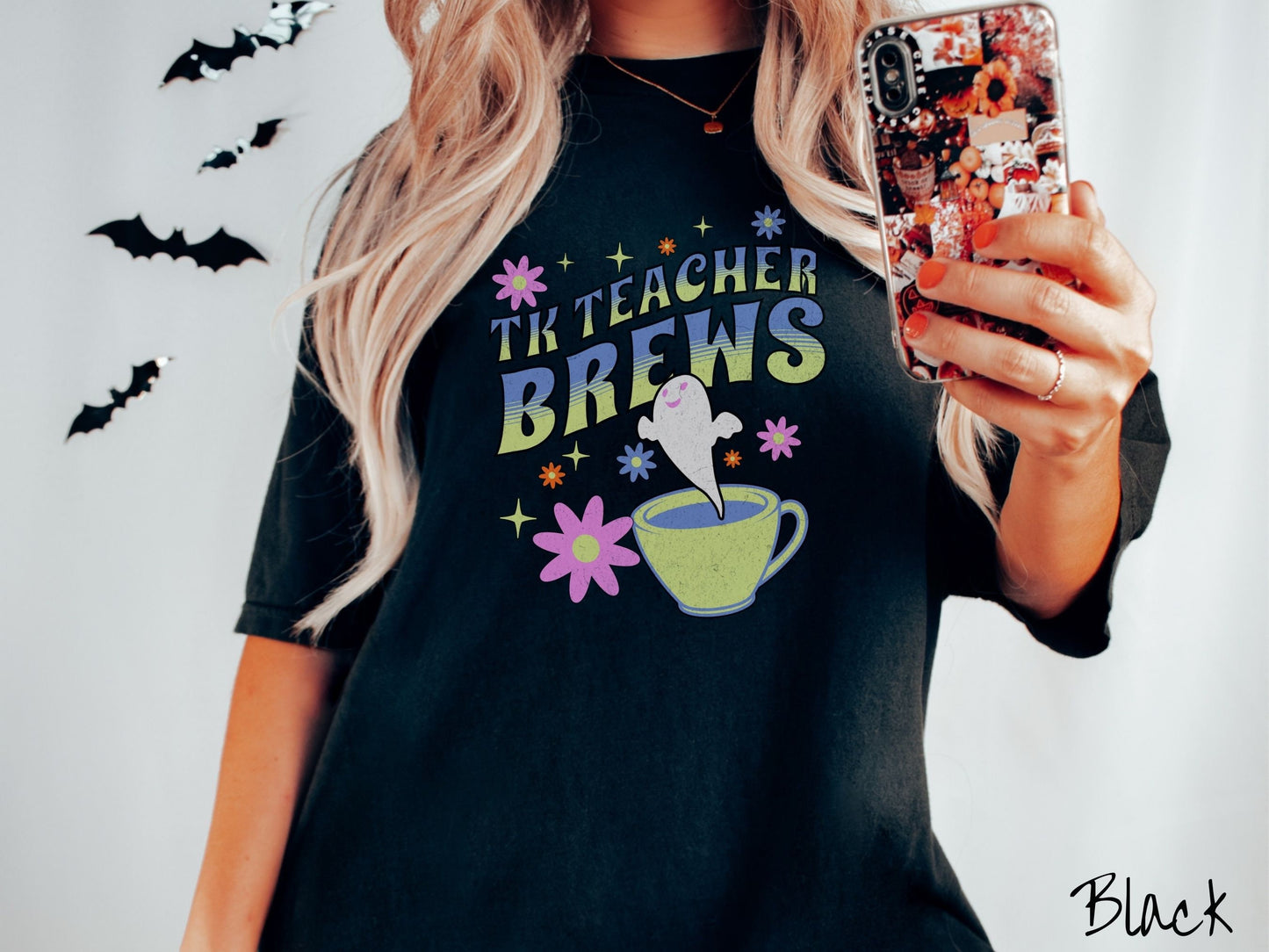 A woman wearing a cute, vintage black colored Comfort Colors t-shirt with the text TK Teacher Brews in blue and green font. Below is a cup of coffee with a smiling ghost floating out of it like hot steam and surrounded by colorful flowers.