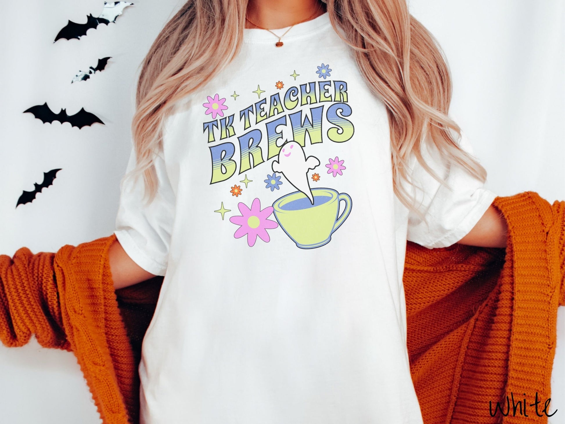 A woman wearing a cute, vintage white colored Comfort Colors t-shirt with the text TK Teacher Brews in blue and green font. Below is a cup of coffee with a smiling ghost floating out of it like hot steam and surrounded by colorful flowers.