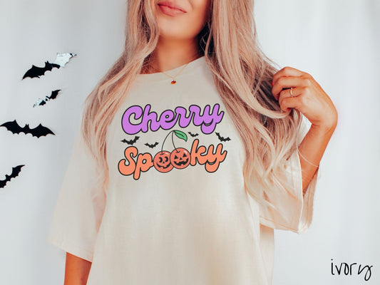 A woman wearing a vintage, ivory colored Comfort Colors t-shirt with the text Cherry Spooky in purple and orange font. The two letter O’s in Spooky are smiling jack o&#39;lantern-themed cherries, and there are black bats flying in the background.