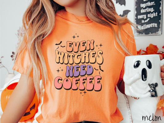 A woman wearing a vintage, melon colored Comfort Colors t-shirt with the text Even Witches Need Coffee in orange, purple, and pink uppercase font. Around the text are black, twinkling stars and black bats flying through the air.