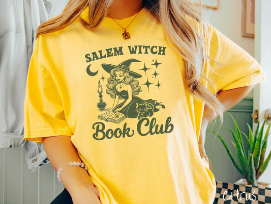 A woman wearing a cute, vintage citrus colored Comfort Colors T-shirt with the text Salem Witch Book Club. In between the text is a young female witch reading a book on her knees surrounded by a candle, black cat, and stars and the moon.