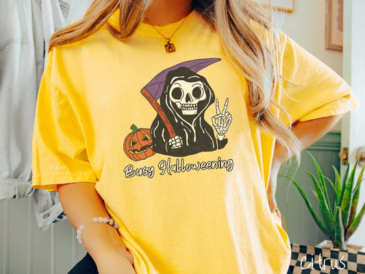 A woman wearing a cute, vintage citrus colored Comfort Colors T-shirt with text Busy Halloweening in white font. Above is a skeleton wearing a black hood holding up a peace sign in one hand and a scythe in the other with a carved pumpkin behind it.