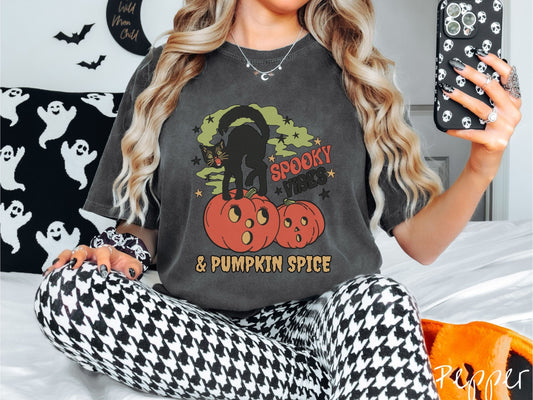 A woman wearing a cute, vintage pepper colored Comfort Colors T-shirt with the text Spooky Vibes & Pumpkin Spice in orange and black font. There is a black cat standing on an orange carved pumpkin and surrounded by green mist and twinkling stars.