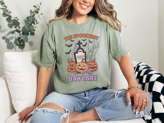 A woman wearing a cute, vintage bay colored Comfort Colors T-shirt with the text The Spookiest Daycare. There are three smiling carved pumpkins sitting in front of a tombstone with a skeleton arm coming out of the ground and black bats in the air.