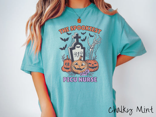 A woman wearing a cute, vintage chalky mint colored Comfort Colors T-shirt with the text The Spookiest PICU Nurse. There are three carved pumpkins sitting in front of a tombstone with a skeleton arm coming out of the ground and black bats in the air.