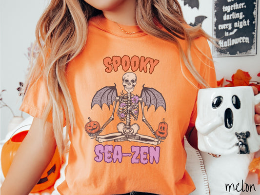 A woman wearing a cute, vintage melon colored Comfort Colors T-shirt with text Spooky Sea-Zen in orange and purple font. There is a skeleton wearing flowers and bat wings sitting in a full lotus yoga pose while holding pumpkins in both hands.