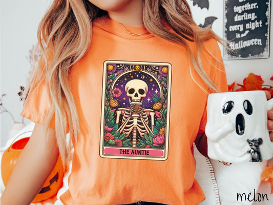 A woman wearing a cute, vintage melon colored Comfort Colors T-shirt with text The Auntie along the bottom of a tarot card. Above is a smiling skeleton holding a to-go cup of coffee up to her mouth, and is surrounded by stars and colorful flowers.