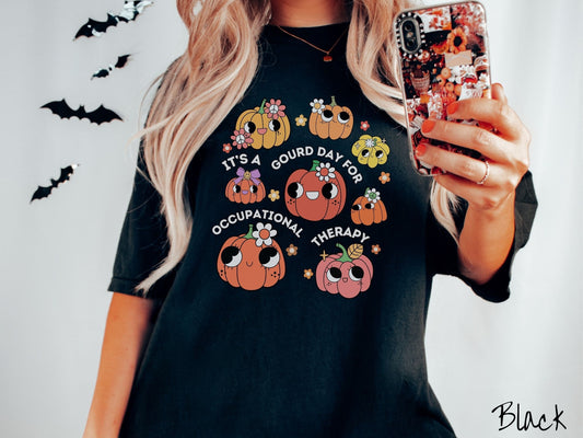 A woman wearing a cute, vintage black colored Comfort Colors T-shirt with text Its a Gourd Day for Occupational Therapy in white font. There is a three by three grid of orange, yellow, and pink pumpkins with girly eyes, wearing flowers and bows.