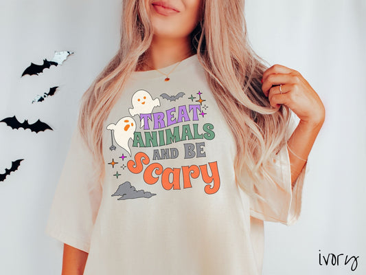 A woman wearing a cute, vintage ivory colored Comfort Colors T-shirt with the text Treat Animals and Be Scary in colorful font. There are orange-eyed white ghosts flying around and black bats and twinkling stars in the sky.