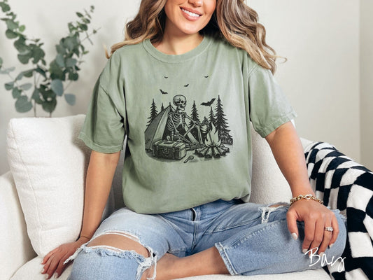 A woman wearing a cute, vintage bay colored Comfort Colors T-shirt with a skeleton sitting in a tent building a fire next to a cooler out in the woods. The campsite is surrounded by evergreen trees and there are bats flying above.