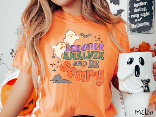 A woman wearing a cute, vintage melon colored Comfort Colors T-shirt with the text Behavior Analyze and Be Scary in colorful font. There are orange-eyed ghosts flying above and a black bat. A black spider is dangling by its web from one ghost.