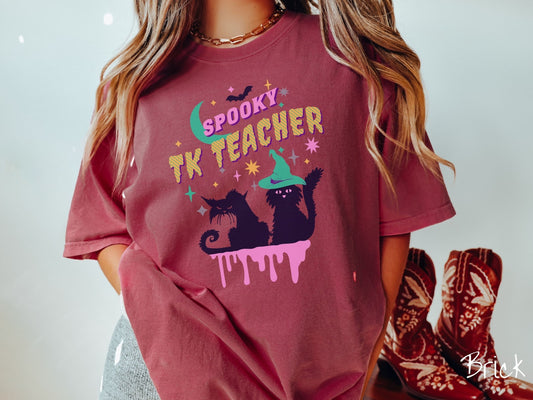 A woman wearing a cute, vintage brick colored Comfort Colors t-shirt with the text Spooky TK Teacher in pink and yellow font. Below are two black cats with pink eyes wearing witch hats sitting beneath colorful stars and a bright green moon.