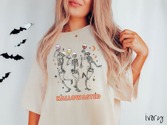 A woman wearing a cute, vintage ivory colored Comfort Colors T-shirt with the text Hallowasted in orange font. Above are three female skeletons dancing while holding wine glasses with different colored wines and surrounded by stars.