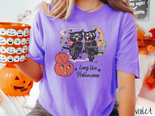 A woman wearing a cute, vintage violet colored Comfort Colors T-shirt with the text Long Live Halloween in black font. Above are two black owls sitting on a tree branch by two scared pumpkins and surrounded by stars and the moon.