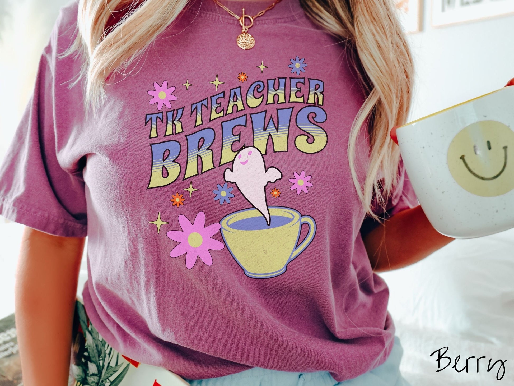 A woman wearing a cute, vintage berry colored Comfort Colors t-shirt with the text TK Teacher Brews in blue and green font. Below is a cup of coffee with a smiling ghost floating out of it like hot steam and surrounded by colorful flowers.