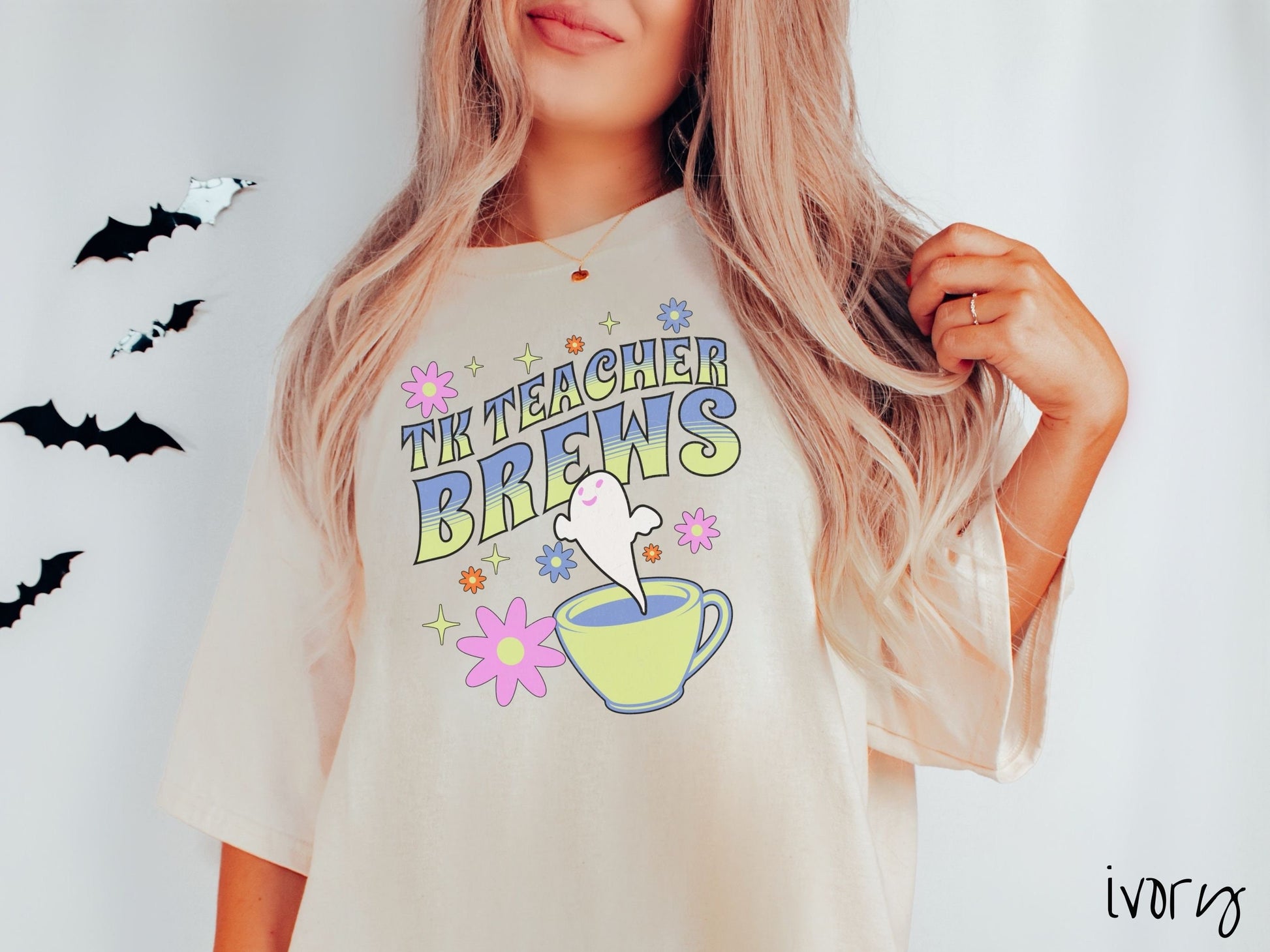 A woman wearing a cute, vintage ivory colored Comfort Colors t-shirt with the text TK Teacher Brews in blue and green font. Below is a cup of coffee with a smiling ghost floating out of it like hot steam and surrounded by colorful flowers.