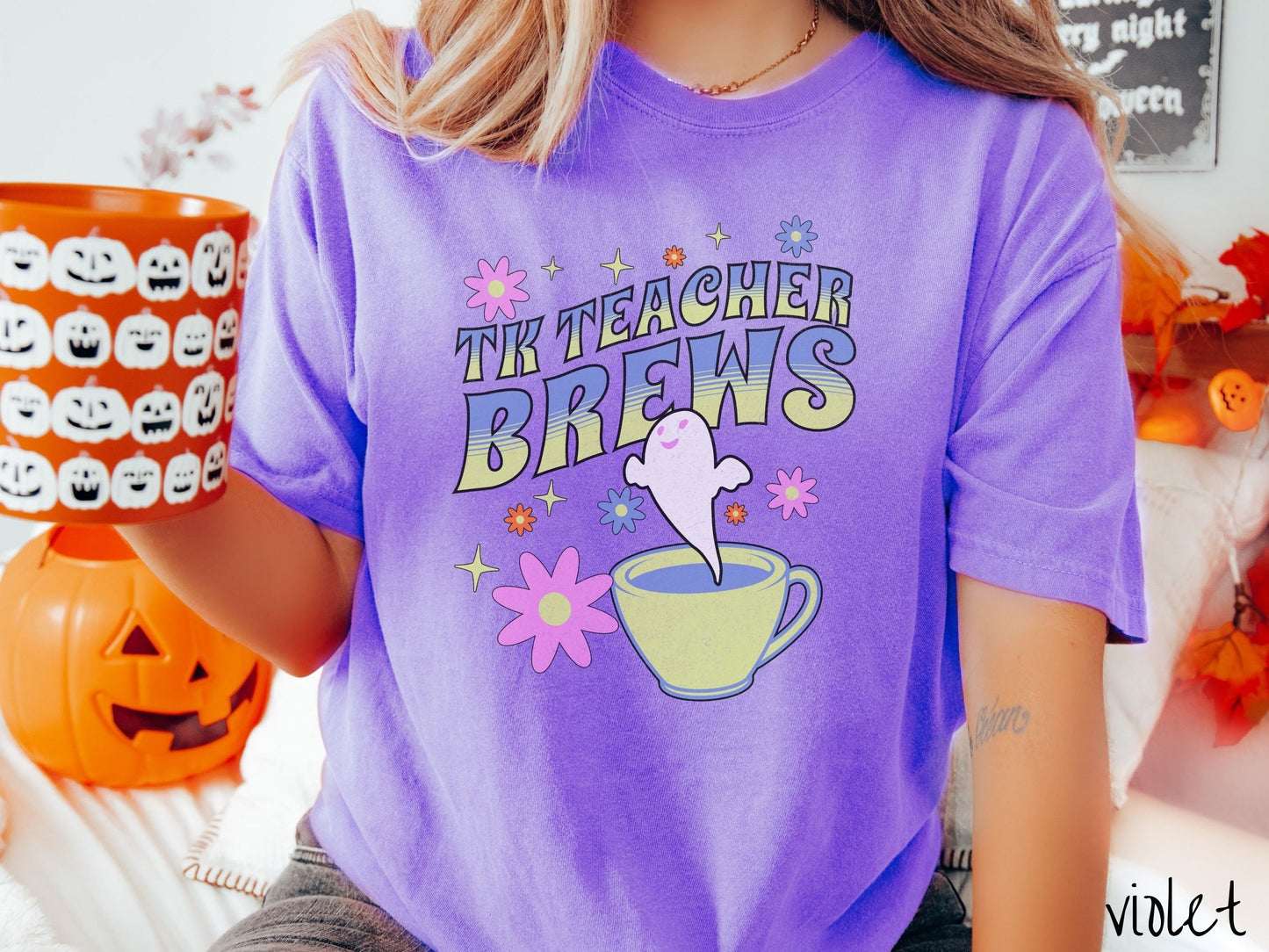 A woman wearing a cute, vintage violet colored Comfort Colors t-shirt with the text TK Teacher Brews in blue and green font. Below is a cup of coffee with a smiling ghost floating out of it like hot steam and surrounded by colorful flowers.