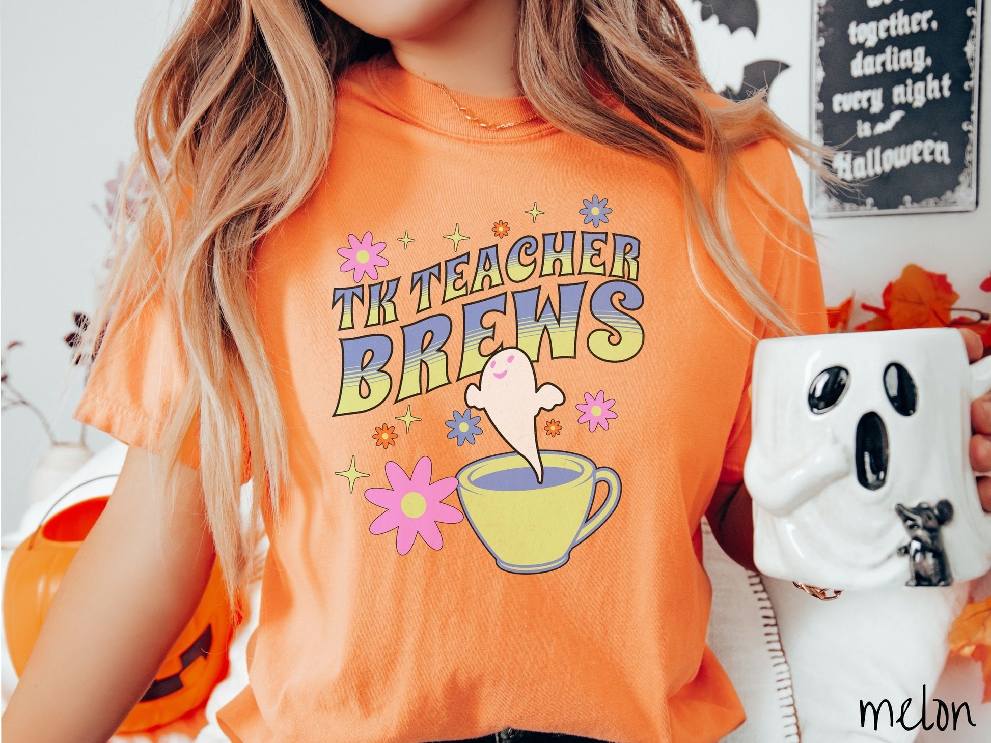 A woman wearing a cute, vintage melon colored Comfort Colors t-shirt with the text TK Teacher Brews in blue and green font. Below is a cup of coffee with a smiling ghost floating out of it like hot steam and surrounded by colorful flowers.