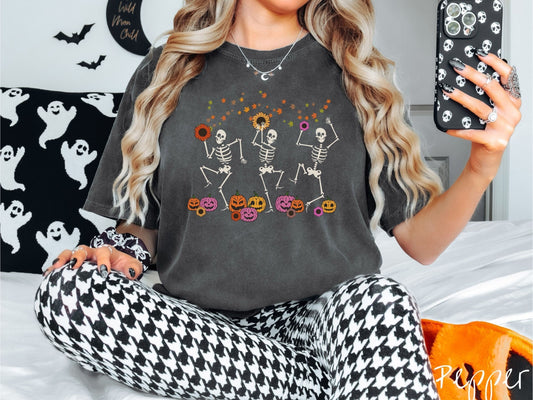 A woman wearing a cute, vintage pepper colored Comfort Colors T-shirt with three white skeletons dancing with hands in the air around orange and pink pumpkins with colorful autumn leaves in the air and colorful sunflowers in their hands.