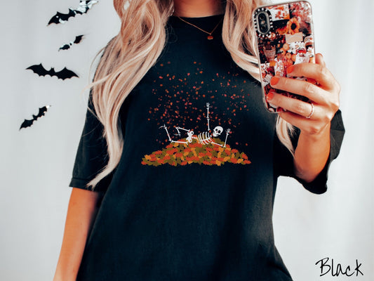 A woman wearing a cute, vintage black colored Comfort Colors T-shirt with a white skeleton playing in a pile of colorful fall leaves. The skeleton is throwing leaves in the air, and there are colorful leaves floating above.