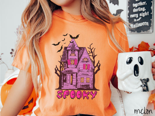 A woman wearing a cute, vintage melon colored Comfort Colors T-shirt with the text Spooky in purple across the bottom. Above is a Purple and pink haunted house with dark trees in the background with black bats flying above.