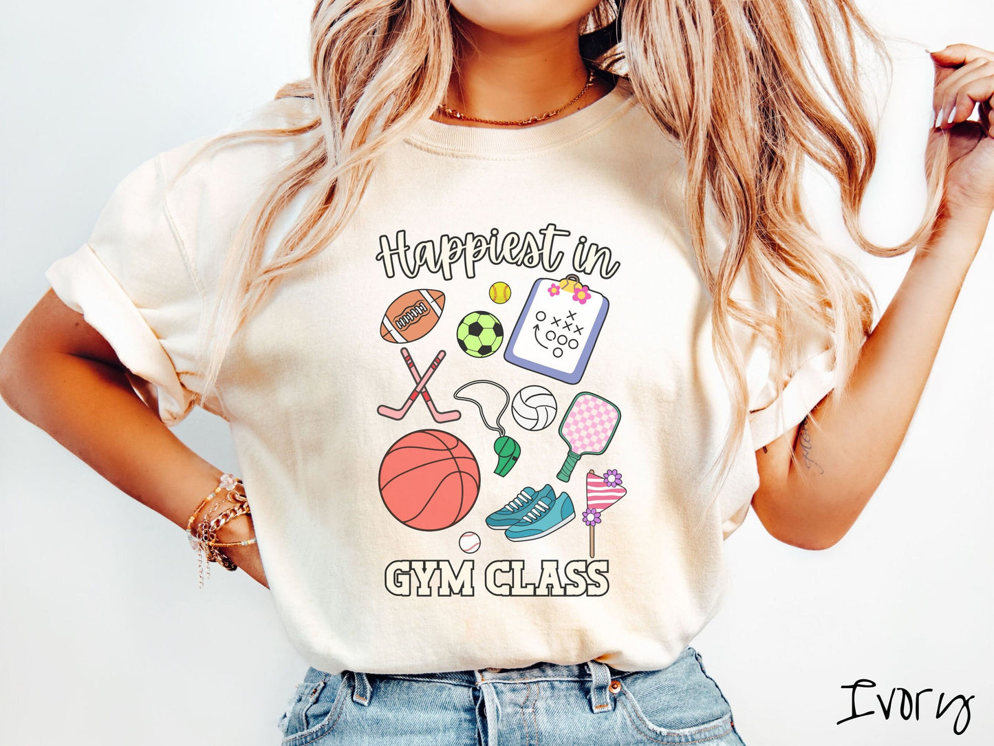 A woman wearing a cute, vintage ivory colored Comfort Colors T-shirt with the text Happiest in Gym Class. In between the text are a football, soccer ball, baseball, hockey sticks, clipboard with plays, basketball, whistle, rackets, and a baseball.