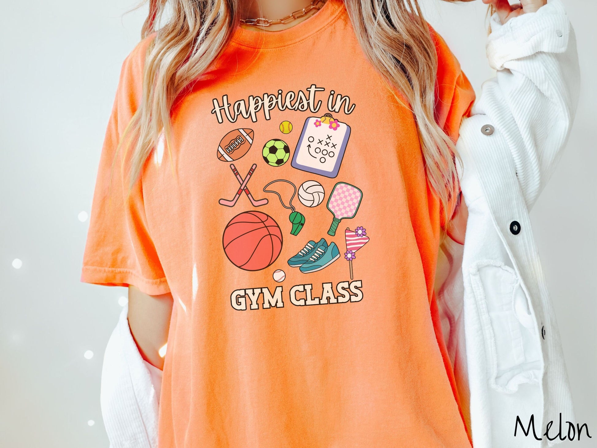 A woman wearing a cute, vintage melon colored Comfort Colors T-shirt with the text Happiest in Gym Class. In between the text are a football, soccer ball, baseball, hockey sticks, clipboard with plays, basketball, whistle, rackets, and a baseball.