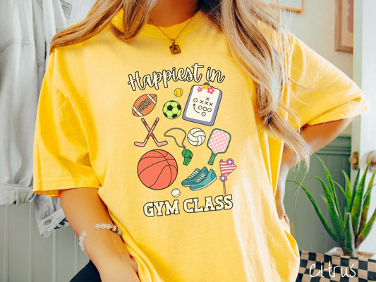 A woman wearing a cute, vintage citrus colored Comfort Colors T-shirt with the text Happiest in Gym Class. In between the text are a football, soccer ball, baseball, hockey sticks, clipboard with plays, basketball, whistle, rackets, and a baseball.