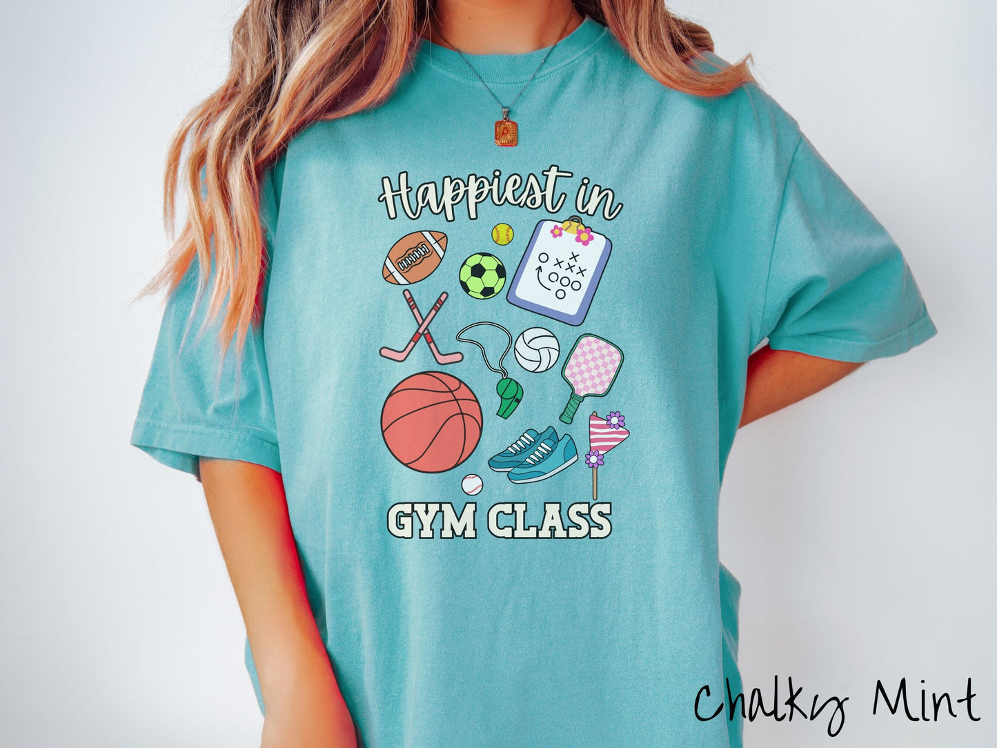 A woman wearing a cute, vintage chalky mint colored Comfort Colors T-shirt with text Happiest in Gym Class. In between the text are a football, soccer ball, baseball, hockey sticks, clipboard with plays, basketball, whistle, rackets, and a baseball.