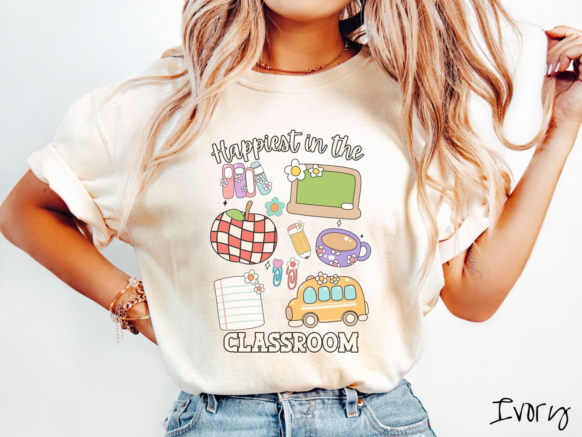 A woman wearing a cute, vintage ivory colored Comfort Colors T-shirt with the text Happiest in the Classroom. In between the text are books, an apple, chalkboard, pencil and paper, coffee cup, yellow school bus, paperclips, and flowers.